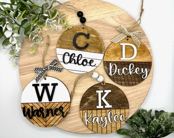 May include: Four round wooden ornaments with a white top half and a brown wood bottom half. Each ornament has a large black letter and a name written in white. The names are Chloe, Dickey, Warner, and Kaylee.