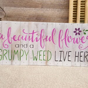May include: A white wooden sign with the text "a beautiful flower and a grumpy weed live here" in pink, green, and black lettering. A purple flower with green leaves is painted on the sign.