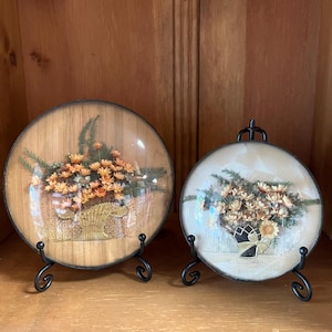May include: Two decorative round wall art pieces. One features a wooden background with a floral arrangement in a basket. The other has a cream background with a similar floral arrangement. Both are displayed on black metal stands.
