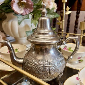 May include: A silver teapot with intricate embossed patterns, a curved spout, and a handle. The teapot sits on small feet and is topped with a decorative finial. Teacups and saucers with floral designs are in the background.