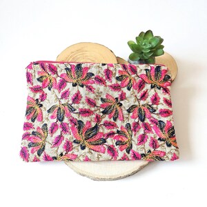 Peut inclure: Pochette rectangulaire avec fermeture éclair rose, ornée d'un motif floral. Le motif présente des fleurs brodées roses, bleu marine et dorées sur un fond beige. La pochette est posée sur une tranche de bois.