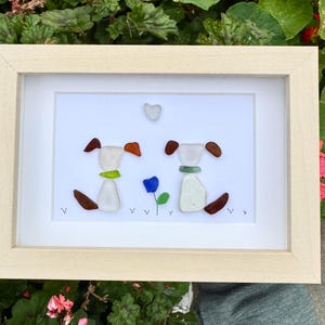 May include: A framed artwork featuring two dog figures crafted from sea glass. The dogs have brown ears and tails, with green and orange accents. A blue flower and a heart-shaped piece of glass are also included in the design.