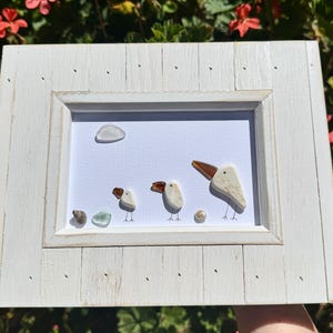 May include: A white wooden frame containing a piece of wall art featuring three birds made from white sea glass and brown glass shards. The birds have thin black ink legs and eyes. A small white glass cloud, a shell, and a green glass piece are included.