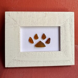 May include: A framed artwork featuring a paw print made of amber-colored sea glass. The sea glass pieces are arranged on a white background within a distressed white wooden frame. The artwork is displayed against a red background.