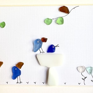 May include: A framed wall art piece featuring sea glass arranged into shapes of birds and flowers on a white background. The composition includes blue, white, and brown sea glass pieces with hand-drawn black ink lines for legs and branches.