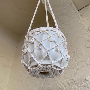 May include: Cream-colored ceramic hanging planter with a macrame net and seashells. The planter is suspended by three cream-colored cords. Decorative pearls are interspersed throughout the macrame. The planter is designed for indoor or outdoor use.