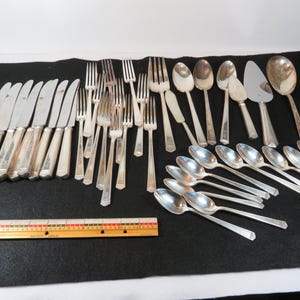 May include: A large collection of silver-toned flatware including knives, forks, and spoons of various sizes, arranged alongside a wooden ruler measuring 12 inches in length, showing the scale of the utensils on a black surface.