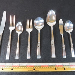 May include: A collection of silver-colored flatware, including knives, forks, and spoons, arranged on a dark surface. The handles feature a decorative design. A wooden ruler with inch measurements is at the bottom.