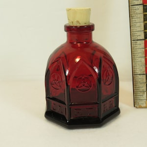 May include: A small, red glass bottle with a cork stopper. The bottle has a gothic design with a repeating pattern of three circles. The bottle is approximately 2 inches tall.