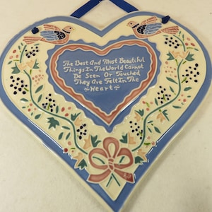 May include: Heart-shaped ceramic wall hanging with a blue border and a cream-colored center. The center features a smaller heart with a quote and is surrounded by floral and bird designs. A blue ribbon is attached for hanging.