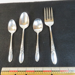 May include: A set of four silver-colored utensils: two spoons, a teaspoon, and a fork. The utensils have a floral design on the handles. The items are arranged on a black surface with a ruler at the bottom.