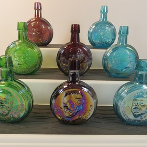 May include: Nine iridescent glass bottles with round bases and portraits of presidents on the front. The bottles are arranged in three rows on a tiered display stand. The bottles are in shades of green, blue, and purple.