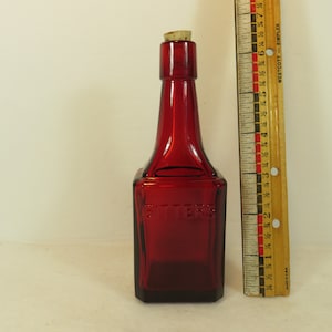 May include: A red glass bottle with a cork stopper. The bottle is square shaped and has the word "Bitters" embossed on the front. The bottle is about 6 inches tall.