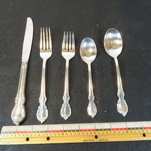 May include: A set of five silver-plated flatware pieces, including a knife, two forks, and two spoons. The pieces are laid out on a black surface with a wooden ruler for scale.