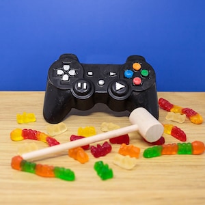 May include: A black video game controller with colorful buttons and a white mallet are on a light wood surface. Scattered around are colorful gummy candies, including worms and bears, against a blue backdrop.