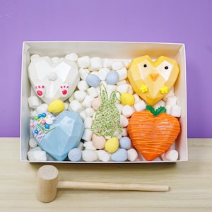 May include: A white box filled with marshmallows, pastel-colored candies, and heart-shaped treats. The treats include a white bear, a yellow chick, a blue heart, and an orange carrot. A wooden mallet rests on the table.