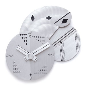 May include: A modern, abstract wall clock with a silver finish and geometric black detailing. The clock features a circular face with diamond and dot patterns, and a unique, layered design. The hands are white with black dots.