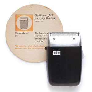 Puede incluir: Una afeitadora eléctrica negra y plateada con el logotipo de Braun en el lateral. La afeitadora está sobre una superficie blanca junto a un posavasos de papel marrón y naranja con texto en alemán.