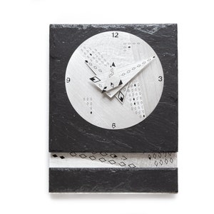 May include: A modern wall clock with a black slate base and a silver-colored clock face. The clock hands and face feature geometric designs. The clock is rectangular with a circular clock face and a decorative lower section.