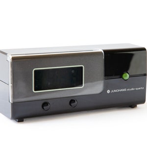 May include: A black and silver digital clock with a green button and the text "JUNGHANS studio-quartz".