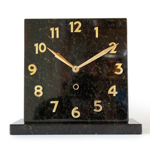 Könnte beinhalten: Eine schwarze Marmoruhr mit goldfarbenen Ziffern und Zeigern. Die Uhr hat ein quadratisches Zifferblatt und einen rechteckigen Sockel.