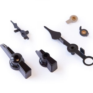 May include: A set of clock hands, including a black hour hand, a black minute hand, and a black second hand. The hands are made of metal and have a black finish. There are also two small metal pieces with a gold finish.