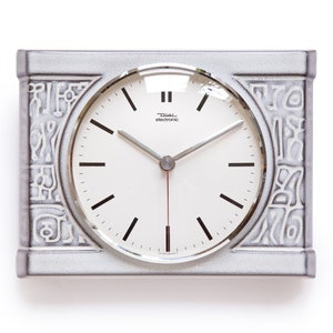 Puede incluir: Un reloj de pared de cerámica blanca con una esfera plateada y agujas negras. El reloj tiene un borde decorativo con un patrón de formas abstractas. La esfera del reloj dice "Tissot Elektrochrono".