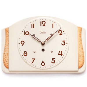 Könnte beinhalten: Eine weiße Keramik-Wanduhr mit braunem Zifferblatt und schwarzen Zeigern. Die Uhr hat römische Ziffern und ein kleines, rundes, schwarzes Zentrum. Die Uhr ist von einem braunen Holzrahmen umgeben.