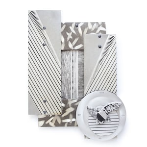 May include: Abstract wall art with a geometric design. The artwork features a combination of silver, gray, and white elements, including vertical stripes, brushstrokes, and a circular element with a patterned design. The piece is mounted on a white background.