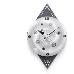 May include: A silver-toned wall clock with a black and gray geometric design. The clock face features black hands, a white inner circle, and decorative tree and dot patterns. The clock is mounted on a black diamond-shaped base.