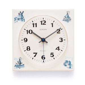 Könnte beinhalten: Eine weiße quadratische Uhr mit schwarzen Zahlen und Zeigern. Das Zifferblatt der Uhr trägt die Aufschrift "EUROPA". Die Uhr ist mit blauen und weißen Illustrationen von Segelbooten und Windmühlen verziert.