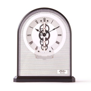 May include: A black and white analogue clock with a visible gear mechanism. The clock has a white face with black Roman numeral hour markers and black hands. The clock is encased in a clear glass dome with a black base. The clock face also has the text "Raliee Quartz".