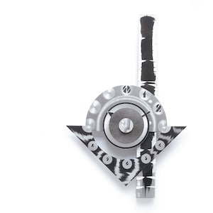 May include: Abstract wall art featuring a geometric design with a circular dial, arrow, and vertical bar. The artwork uses a black and white color scheme with textured patterns and metallic accents. The piece is mounted on a white background.
