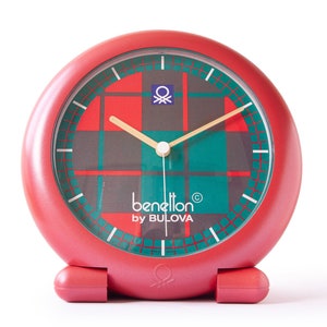 Puede incluir: Un reloj despertador rojo con una cara de cuadros verdes y rojos. La esfera del reloj tiene el texto "benetton® by BULOVA" y un pequeño logotipo.