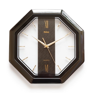 Könnte beinhalten: Schwarze achteckige Wanduhr mit goldenen römischen Ziffern und weißem Zifferblatt. Die Uhr hat ein schwarzes und goldenes Band mit dem Wort "Mebus" und "QUARTZ" darauf gedruckt.