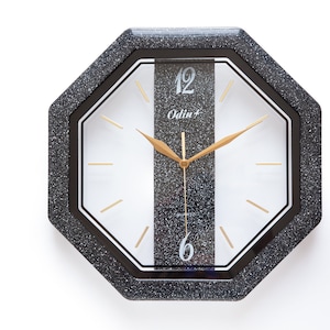Könnte beinhalten: Eine schwarze achteckige Wanduhr mit weißem Zifferblatt und goldenen Zeigern. Die Uhr hat eine silberne Glitzer-Oberfläche und der Text "Odin+" ist auf dem Zifferblatt aufgedruckt.
