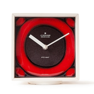 Peut inclure: Horloge carrée Junghans electronic ATO-MAT avec un cadran rouge et noir. L'horloge a un cadre argenté et des aiguilles blanches. Le cadran porte le logo Junghans et les mots "ATO-MAT". L'horloge est sur une base blanche.