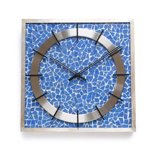 Könnte beinhalten: Quadratische Wanduhr mit einem blauen Mosaikdesign und einem silbernen Metallring um das Zifferblatt. Die Uhrzeiger sind schwarz.