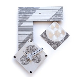 May include: Abstract wall clock with a geometric design. The clock features a white face with black numbers and hands, and a gray and white striped frame. The clock has a unique, modern aesthetic.