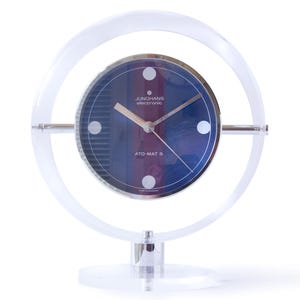 May include: A vintage Junghans ATO-MAT S clock with a clear acrylic frame. The clock face is dark blue with silver hands and markers. The clock has a circular design and sits on a clear base.