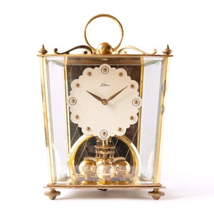 May include: A gold-toned, square-shaped clock with a glass face and a pendulum. The clock face has a white background with black Roman numeral hour markers and black hands. The clock is on a white background.