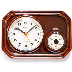 Puede incluir: Un reloj de madera marrón con una esfera blanca y agujas negras. El reloj tiene un temporizador redondo más pequeño con una esfera blanca y agujas negras. La esfera del reloj dice "KIENZLE QUARTZ".