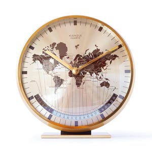May include: A round Kienzle Quartz desk clock with a gold-toned frame and a metallic face featuring a dark brown world map. The clock includes gold hour and minute hands, black hour markers, and a curved 24-hour scale at the bottom of the dial.