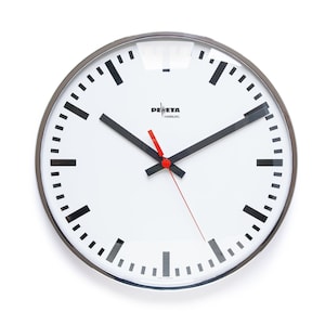 May include: A white analog clock with black hour and minute hands and a red second hand. The clock face has black markings for the hours and minutes. The clock is surrounded by a silver metal frame. The clock face also has the text "PELETA HAMBURG" printed on it.