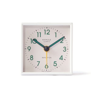 Könnte beinhalten: Weißer quadratischer Wecker mit cremefarbenem Zifferblatt und schwarzen Zeigern. Das Zifferblatt hat schwarze Zahlen und grüne Akzente. Die Uhr zeigt "Kienzle Quartz" und "Repetition" auf dem Zifferblatt an. Die Uhr ist in Deutschland hergestellt.