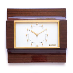 May include: A rectangular wall clock featuring a dark brown polished wood frame with a white face, gold-toned hands, and gold-toned circular hour markers. The clock face includes black minute indices and the text Junghans Quartz in the corner.