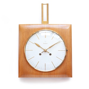 May include: A square wall clock with a light brown wood frame and a white circular face. The clock features gold-colored hands, thin gold hour markers, and a gold metal hanging bracket extending from the top. The brand name Junghans is printed in small black text on the white face.