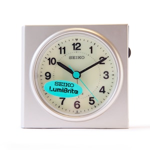 Könnte beinhalten: Ein silberner quadratischer Wecker mit weißem Zifferblatt und schwarzen Zeigern. Die Uhr hat einen blau-weißen Aufkleber mit der Aufschrift "SEIKO LumiBrite".