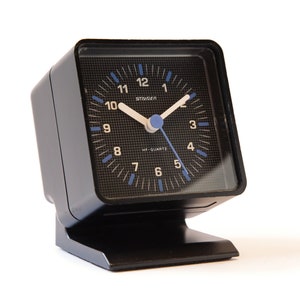 May include: A black square alarm clock with a white face and blue hands. The clock has a black base and says "Staiger" and "HF-Quartz" on the face.