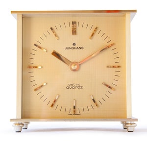 May include: A gold-toned Junghans Astra Quartz clock with a square face and black hands. The clock has a white background with black Roman numeral hour markers and black minute markers.